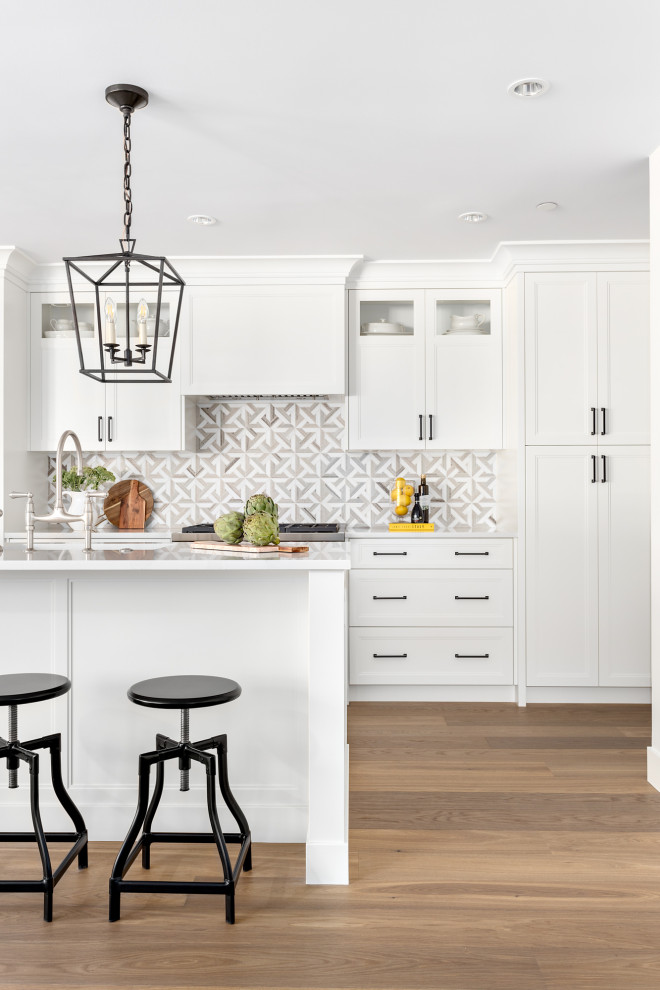 This is an example of a mid-sized transitional single-wall open plan kitchen in Vancouver with a farmhouse sink, recessed-panel cabinets, white cabinets, quartz benchtops, beige splashback, mosaic tile splashback, panelled appliances, medium hardwood floors, with island and white benchtop.
