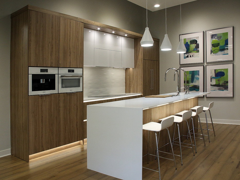 This is an example of a large modern single-wall open plan kitchen in Grand Rapids with an undermount sink, flat-panel cabinets, light wood cabinets, quartz benchtops, white splashback, porcelain splashback, panelled appliances, medium hardwood floors, with island, brown floor and white benchtop.