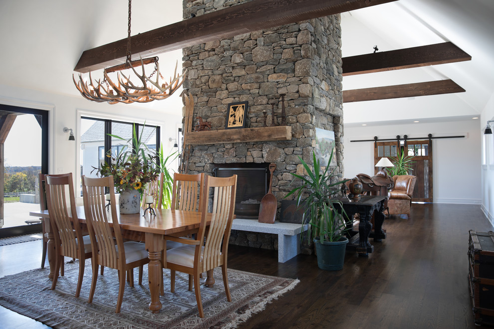 Inspiration for a country open plan dining in New York with white walls, dark hardwood floors, a two-sided fireplace, a stone fireplace surround and brown floor.