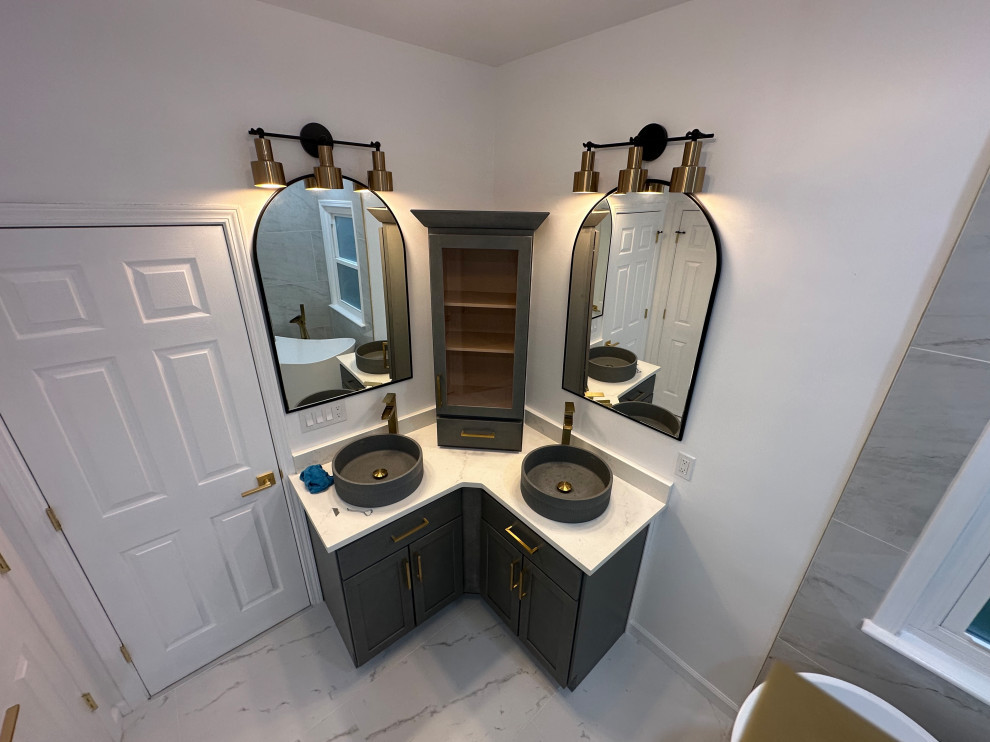 Corner double vanity with gray cabinets, vessel sinks, arched mirrors, brass lighting, and central storage cabinet.