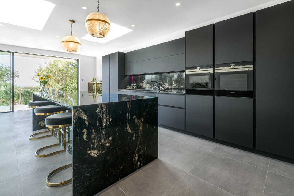 Large contemporary l-shaped open plan kitchen in Sussex with an integrated sink, flat-panel cabinets, black cabinets, granite benchtops, black splashback, granite splashback, black appliances, with island, grey floor and black benchtop.