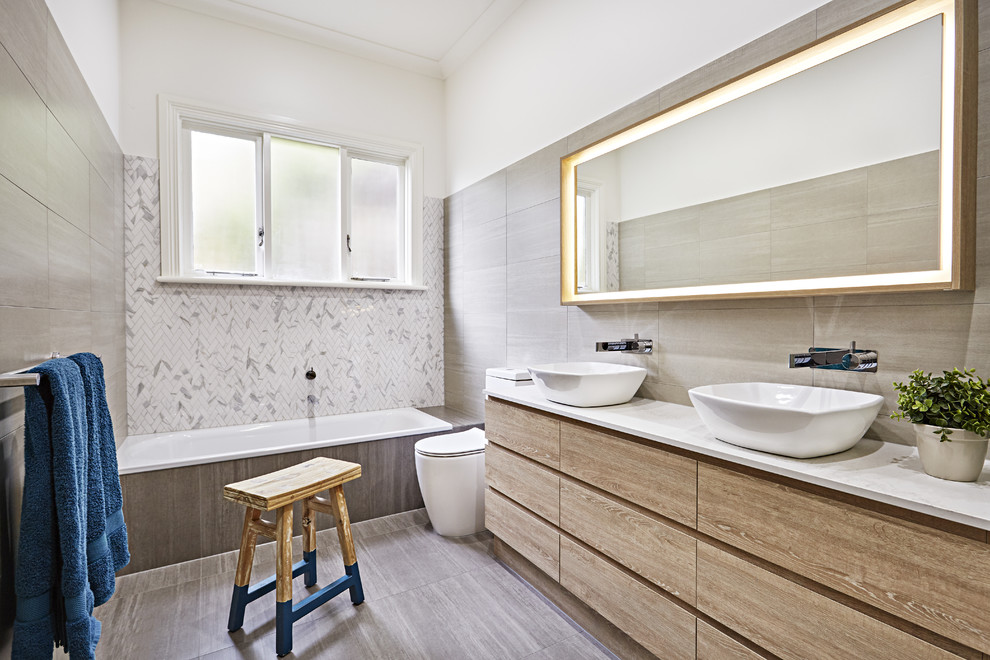 Design ideas for a mid-sized contemporary master bathroom in Melbourne with marble benchtops, flat-panel cabinets, light wood cabinets, a drop-in tub, a one-piece toilet, brown tile, gray tile, white tile, white walls, a vessel sink and brown floor.
