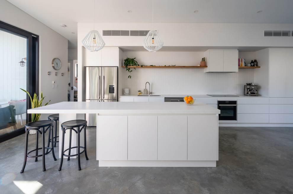 Modern open plan kitchen in Perth with white cabinets, white splashback, ceramic splashback, stainless steel appliances, concrete floors, with island, grey floor and white benchtop.