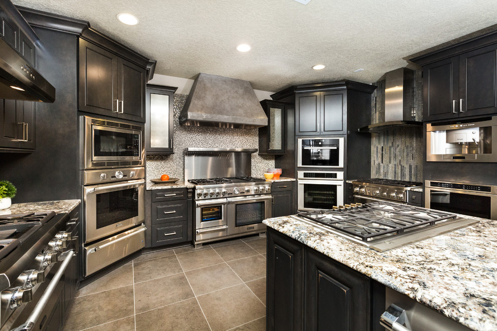 This is an example of a large transitional u-shaped eat-in kitchen in Albuquerque with granite benchtops, glass tile splashback, stainless steel appliances, with island, recessed-panel cabinets, black cabinets, grey splashback and travertine floors.