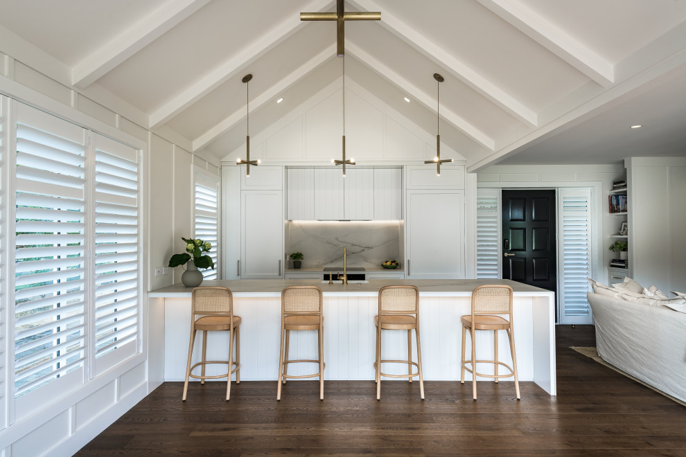 Design ideas for a mid-sized traditional single-wall open plan kitchen in Christchurch with a farmhouse sink, shaker cabinets, white cabinets, white splashback, panelled appliances, medium hardwood floors, with island, brown floor, white benchtop and exposed beam.