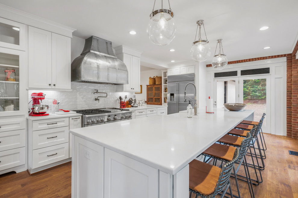 Design ideas for a traditional kitchen in Raleigh with a farmhouse sink, shaker cabinets, white cabinets, white splashback, subway tile splashback, stainless steel appliances, medium hardwood floors, with island and white benchtop.