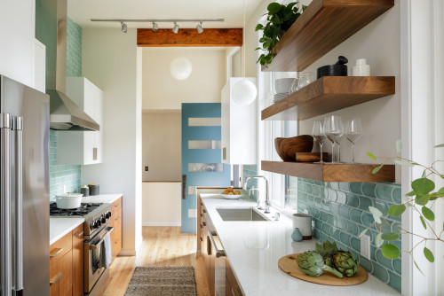 Narrow modern kitchen with wood kitchen cabinets, white countertops, teal backsplash tiles, and open wooden shelves styled with plants and glassware.
