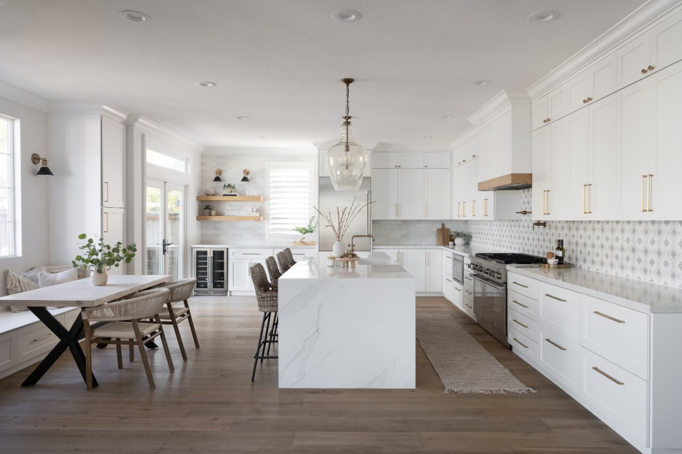 Photo of a large transitional l-shaped eat-in kitchen in San Diego with a farmhouse sink, shaker cabinets, white cabinets, quartz benchtops, white splashback, ceramic splashback, stainless steel appliances, medium hardwood floors, with island, brown floor and white benchtop.