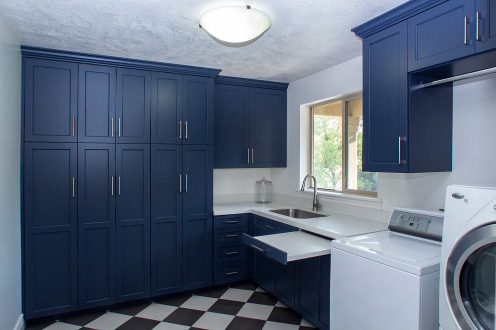 This is an example of a large transitional l-shaped utility room in Salt Lake City with an undermount sink, shaker cabinets, blue cabinets, quartz benchtops, white splashback, engineered quartz splashback, white walls, ceramic floors, a side-by-side washer and dryer, multi-coloured floor and white benchtop.