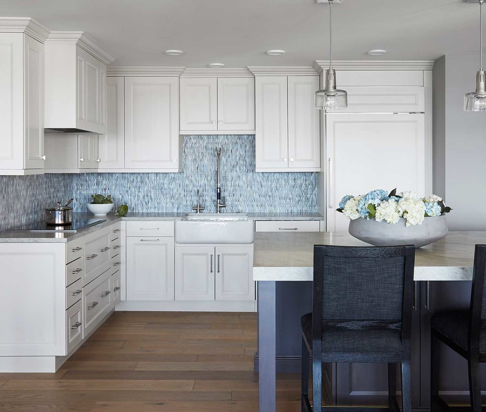 Large contemporary u-shaped open plan kitchen in Chicago with a farmhouse sink, recessed-panel cabinets, white cabinets, quartzite benchtops, blue splashback, glass tile splashback, panelled appliances, light hardwood floors, with island, brown floor and white benchtop.