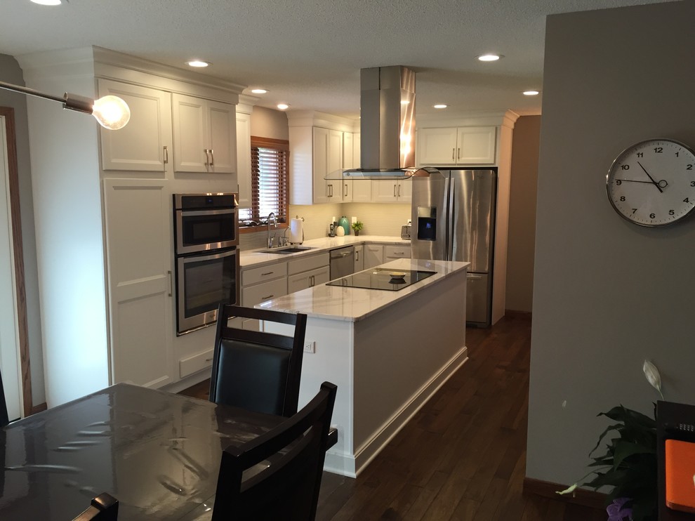 Example of a large transitional l-shaped dark wood floor home design design with an undermount sink, flat-panel cabinets, white cabinets, quartz countertops, white backsplash, ceramic backsplash, stainless steel appliances and an island