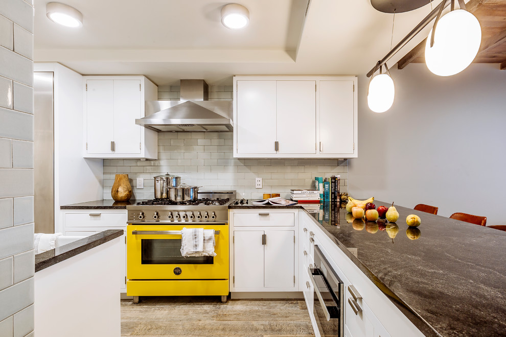 This is an example of a small contemporary u-shaped separate kitchen in New York with a farmhouse sink, flat-panel cabinets, white cabinets, grey splashback, subway tile splashback, coloured appliances, a peninsula, granite benchtops, light hardwood floors, beige floor and wood.