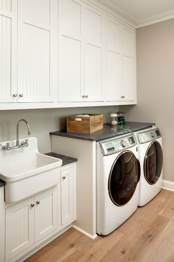 Photo of a mid-sized beach style galley dedicated laundry room in Minneapolis with a farmhouse sink, white cabinets, soapstone benchtops, beige walls, light hardwood floors, a side-by-side washer and dryer and recessed-panel cabinets.