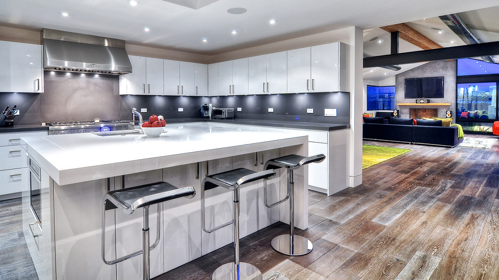 Large modern u-shaped eat-in kitchen in Orange County with a drop-in sink, flat-panel cabinets, white cabinets, glass benchtops, black splashback, stone tile splashback, stainless steel appliances, dark hardwood floors and with island.