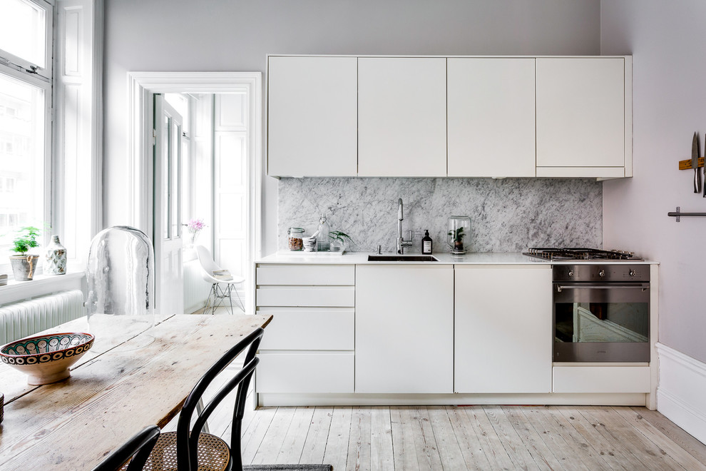 Scandinavian single-wall kitchen in Stockholm with a single-bowl sink, white cabinets, marble benchtops, marble splashback, white appliances, light hardwood floors and no island.