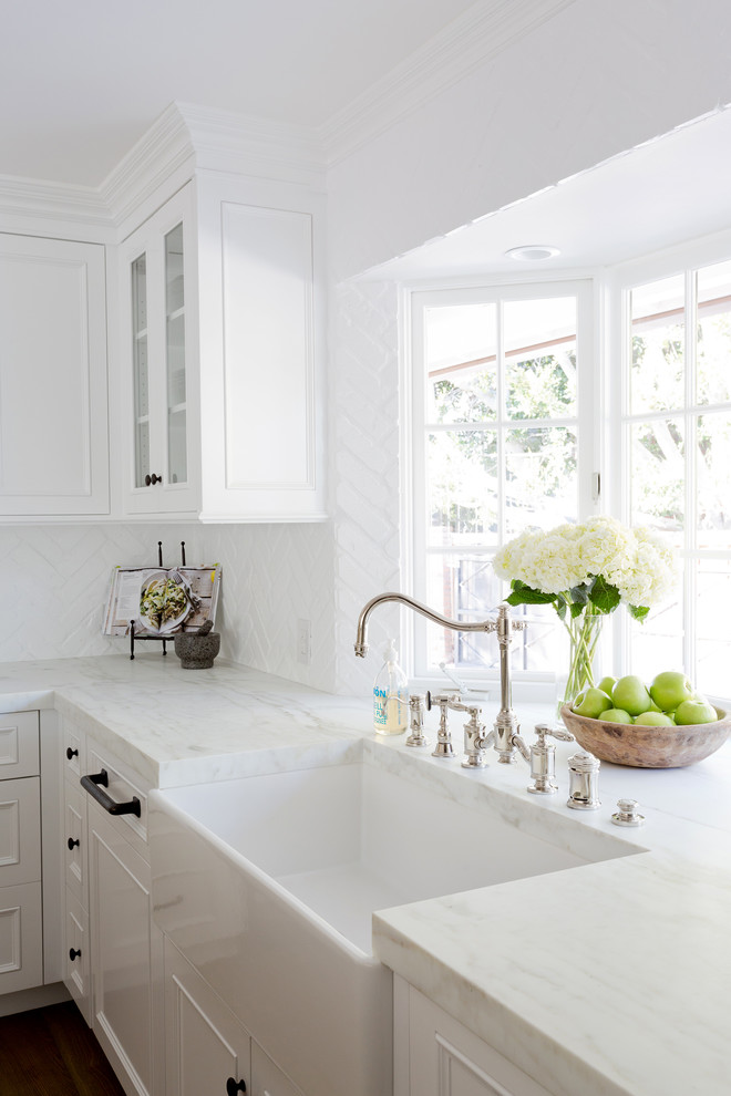Example of a large transitional l-shaped light wood floor open concept kitchen design in Orange County with a farmhouse sink, shaker cabinets, white cabinets, marble countertops, white backsplash, brick backsplash, stainless steel appliances and an island