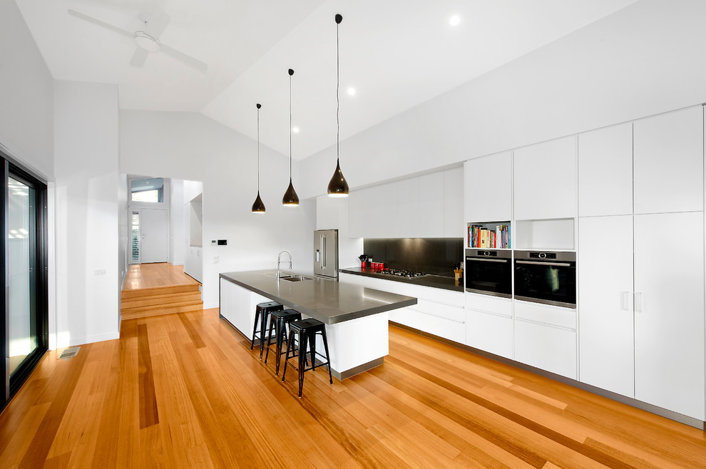 Design ideas for a large contemporary galley open plan kitchen in Melbourne with a double-bowl sink, flat-panel cabinets, white cabinets, quartz benchtops, black splashback, stone slab splashback, stainless steel appliances, light hardwood floors and with island.