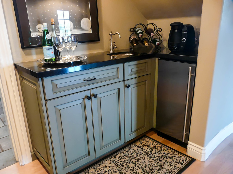 This is an example of a small transitional l-shaped wet bar in San Diego with an undermount sink, raised-panel cabinets, green cabinets, granite benchtops and light hardwood floors.