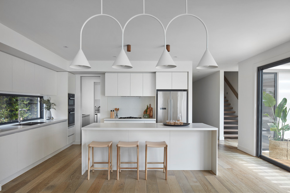 Inspiration for a contemporary l-shaped open plan kitchen in Melbourne with a double-bowl sink, flat-panel cabinets, white cabinets, white splashback, stainless steel appliances, light hardwood floors, with island and brown floor.