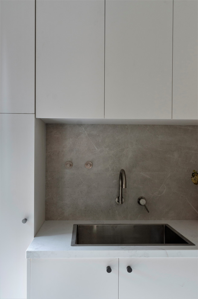 Design ideas for a small modern l-shaped dedicated laundry room in Melbourne with a single-bowl sink, flat-panel cabinets, beige cabinets, quartz benchtops, grey splashback, porcelain splashback, grey walls, porcelain floors, a side-by-side washer and dryer, grey floor and white benchtop.