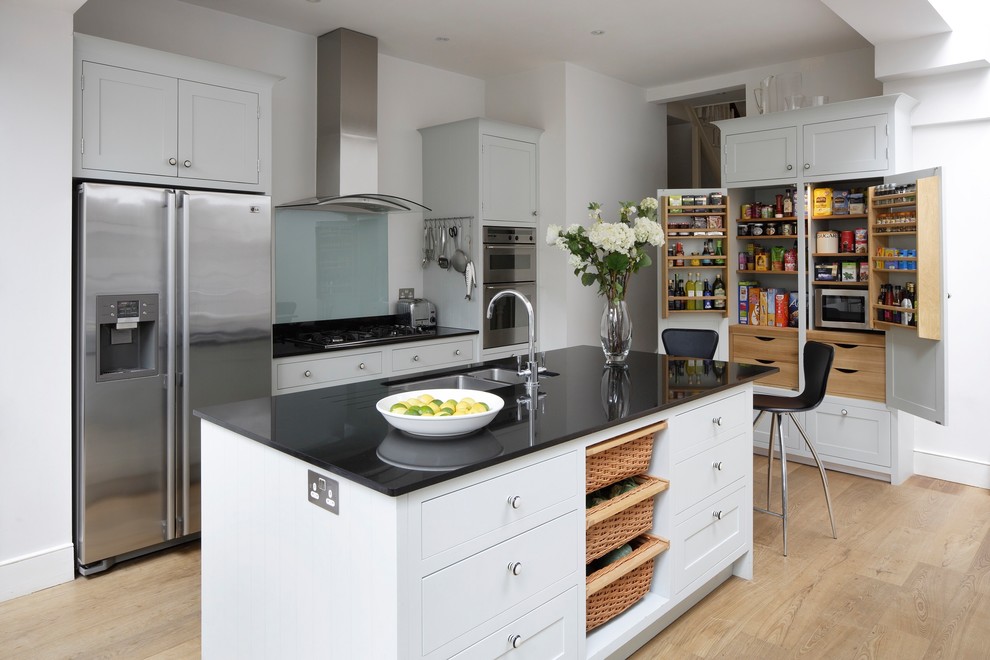 Mid-sized transitional single-wall open plan kitchen in London with a drop-in sink, shaker cabinets, white cabinets, granite benchtops, glass sheet splashback, stainless steel appliances, medium hardwood floors, with island and black benchtop.