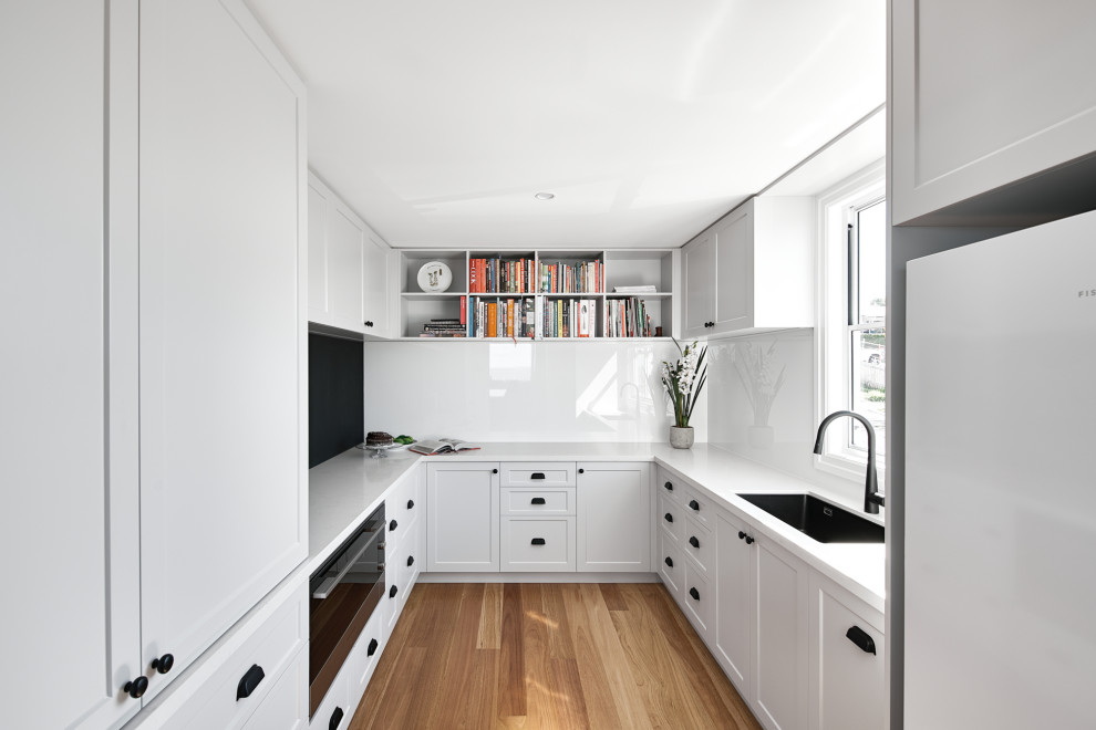 Photo of a mid-sized beach style u-shaped kitchen in Brisbane with an undermount sink, shaker cabinets, white cabinets, white splashback, black appliances, with island, brown floor and beige benchtop.