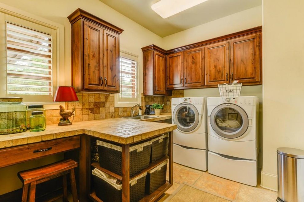 Large mediterranean l-shaped dedicated laundry room in Austin with an utility sink, raised-panel cabinets, medium wood cabinets, tile benchtops, beige walls, ceramic floors, a side-by-side washer and dryer, beige floor and beige benchtop.
