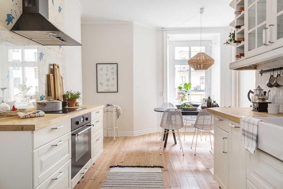 This is an example of a scandinavian galley eat-in kitchen in Gothenburg with a farmhouse sink, raised-panel cabinets, white cabinets, white splashback, subway tile splashback, black appliances, light hardwood floors and no island.