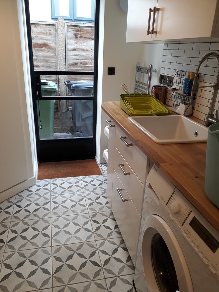 Photo of a small contemporary galley laundry cupboard in London with a farmhouse sink, wood benchtops, white walls, ceramic floors, an integrated washer and dryer, grey floor and multi-coloured benchtop.