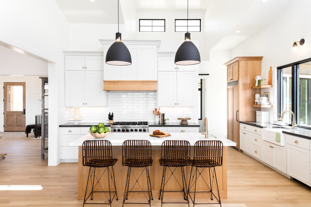 Eat-in kitchen - large cottage l-shaped light wood floor and beige floor eat-in kitchen idea in Salt Lake City with a farmhouse sink, shaker cabinets, white backsplash, ceramic backsplash, black countertops, granite countertops, white cabinets, paneled appliances and an island