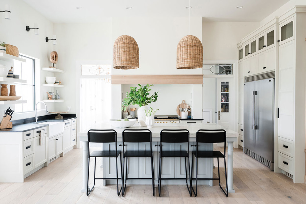 This is an example of a mid-sized beach style u-shaped open plan kitchen in Salt Lake City with a farmhouse sink, shaker cabinets, white cabinets, stainless steel appliances, light hardwood floors, with island, beige floor and black benchtop.