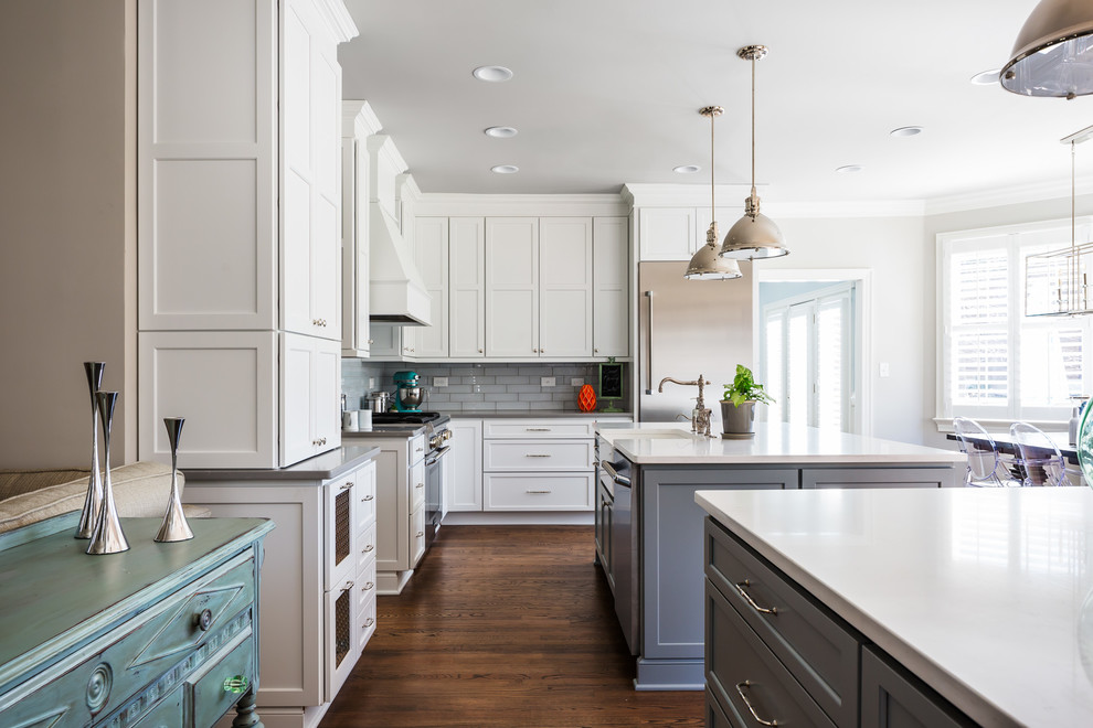 This is an example of an expansive transitional l-shaped open plan kitchen in Raleigh with a farmhouse sink, white cabinets, quartz benchtops, grey splashback, stainless steel appliances, medium hardwood floors, multiple islands, recessed-panel cabinets, glass tile splashback and brown floor.
