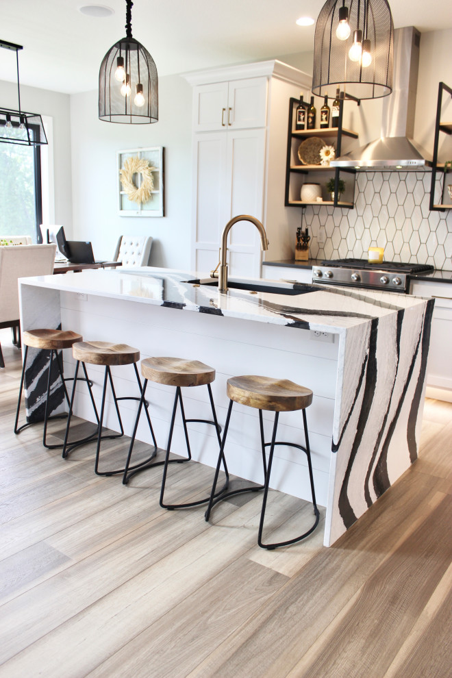 This is an example of a mid-sized transitional l-shaped eat-in kitchen in Other with a farmhouse sink, recessed-panel cabinets, white cabinets, quartz benchtops, white splashback, ceramic splashback, stainless steel appliances, medium hardwood floors, with island, brown floor and multi-coloured benchtop.