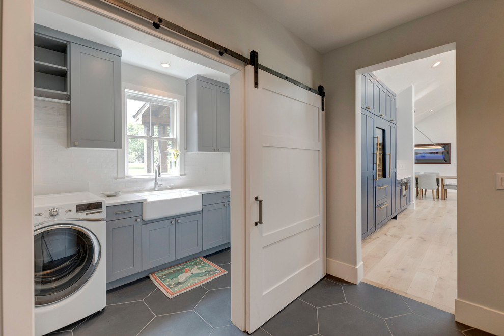 This is an example of a country single-wall dedicated laundry room in Denver with a farmhouse sink, shaker cabinets, grey cabinets, white walls, a side-by-side washer and dryer, grey floor and white benchtop.