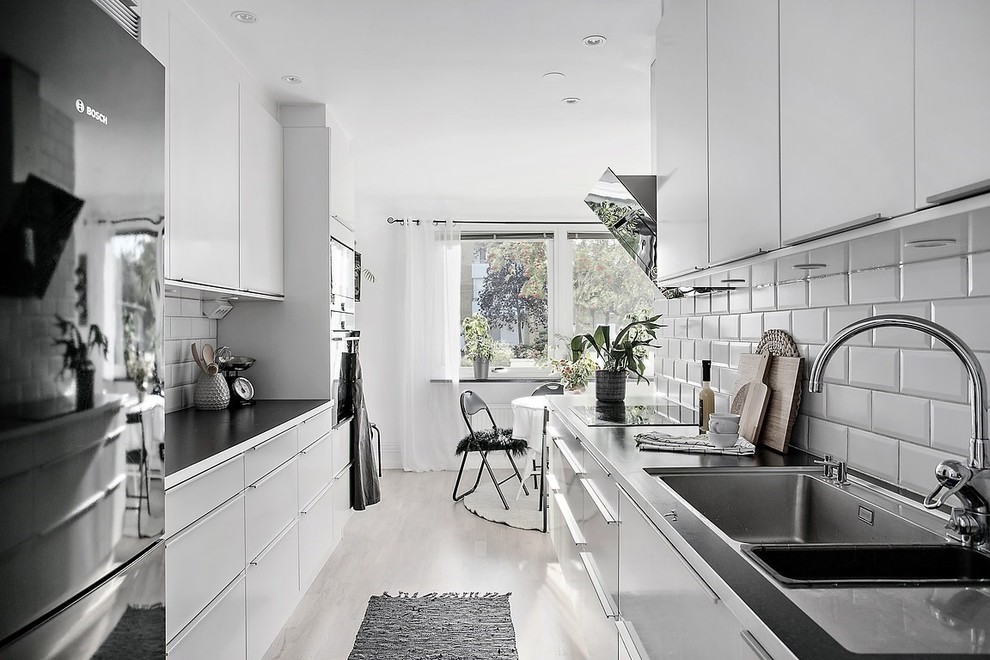 This is an example of a scandinavian galley eat-in kitchen in Gothenburg with a double-bowl sink, flat-panel cabinets, white cabinets, white splashback, subway tile splashback, stainless steel appliances, light hardwood floors and no island.