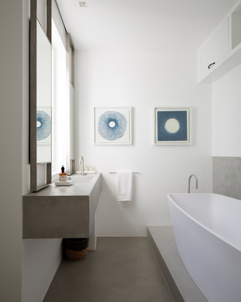 Inspiration for a mid-sized contemporary master bathroom in London with flat-panel cabinets, white cabinets, a freestanding tub, a wall-mount toilet, gray tile, white walls, concrete floors, an integrated sink, concrete benchtops and grey floor.