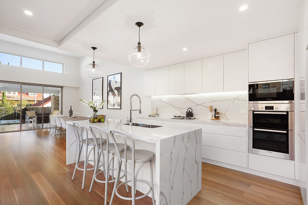 Inspiration for a contemporary galley open plan kitchen in Sydney with quartz benchtops, with island, a double-bowl sink, flat-panel cabinets, white cabinets, multi-coloured splashback, stone slab splashback, stainless steel appliances and medium hardwood floors.