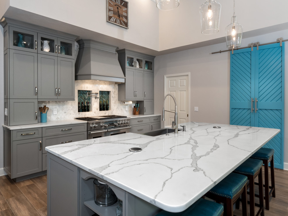 Large transitional l-shaped eat-in kitchen in Miami with a farmhouse sink, shaker cabinets, grey cabinets, quartz benchtops, grey splashback, glass sheet splashback, stainless steel appliances, ceramic floors, with island, brown floor and white benchtop.