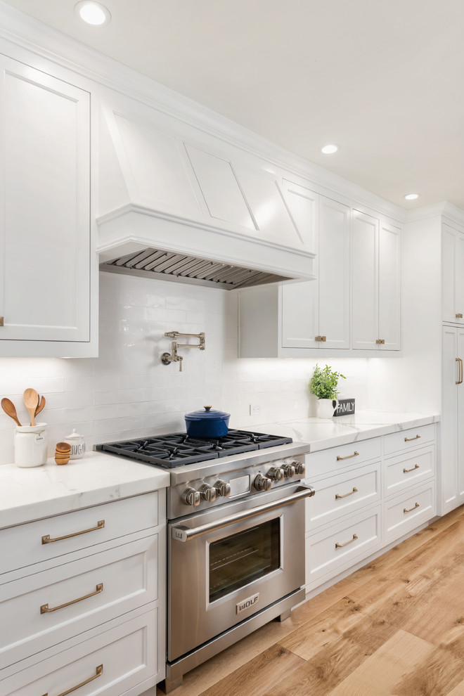 This is an example of a large country u-shaped open plan kitchen in San Francisco with a farmhouse sink, white cabinets, marble benchtops, white splashback, ceramic splashback, panelled appliances, medium hardwood floors, with island, brown floor, white benchtop and recessed-panel cabinets.