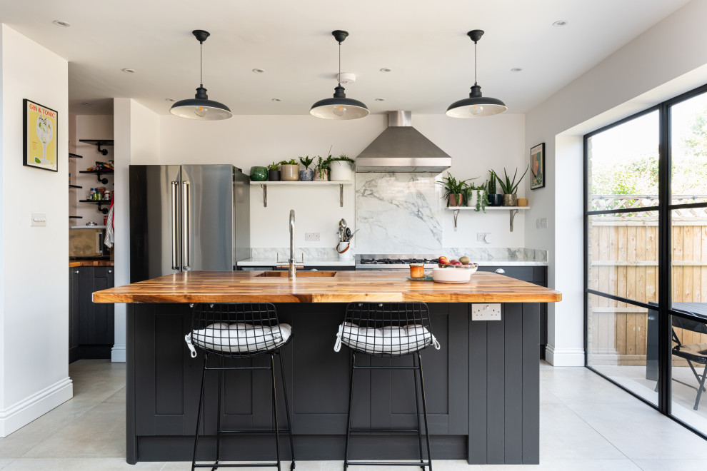 Cette image montre une cuisine design avec des portes de placard noires, un plan de travail en bois, une crédence blanche, une crédence en marbre, une péninsule et un sol gris.