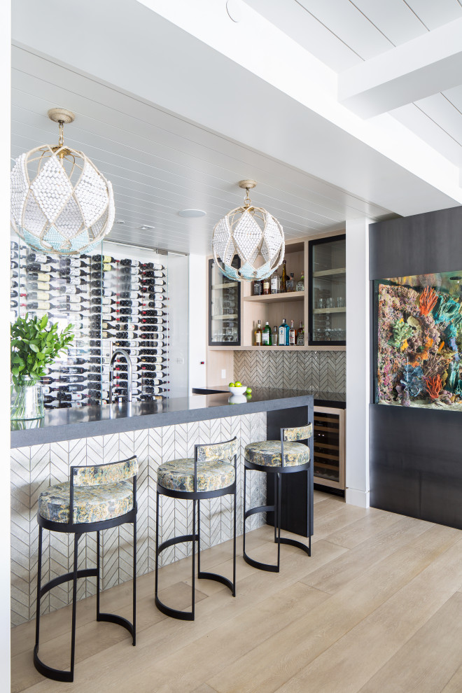 This is an example of a large beach style l-shaped seated home bar in Orange County with glass-front cabinets, light wood cabinets, grey splashback, light hardwood floors, mosaic tile splashback, beige floor and grey benchtop.
