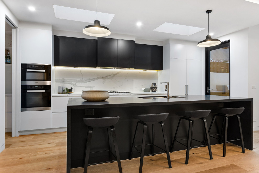 This is an example of a large contemporary galley kitchen in Melbourne with a double-bowl sink, flat-panel cabinets, black cabinets, quartz benchtops, marble splashback, stainless steel appliances, with island, brown floor, black benchtop, white splashback and medium hardwood floors.