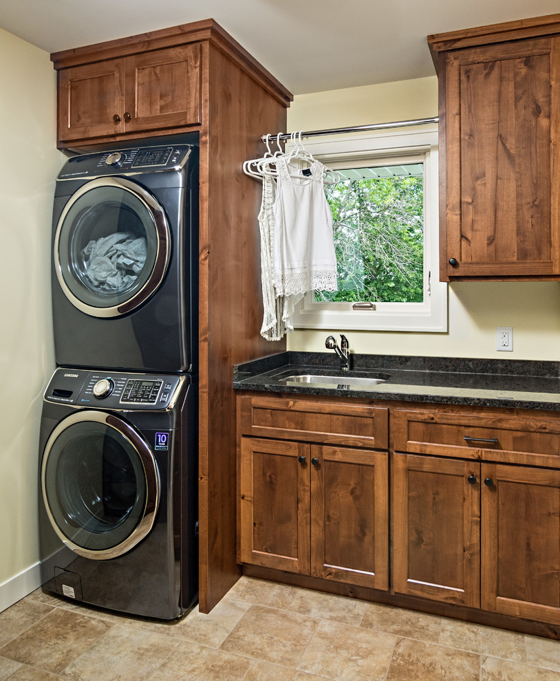 Inspiration for a mid-sized arts and crafts single-wall dedicated laundry room in Minneapolis with an undermount sink, shaker cabinets, medium wood cabinets, granite benchtops, beige walls, porcelain floors, a stacked washer and dryer, beige floor and black benchtop.