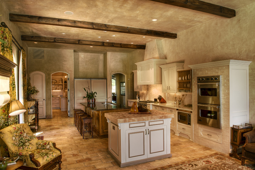 This is an example of a mid-sized mediterranean galley open plan kitchen in Other with panelled appliances, wood benchtops, brick floors, a double-bowl sink, beaded inset cabinets, white cabinets, beige splashback, travertine splashback, multiple islands and beige floor.