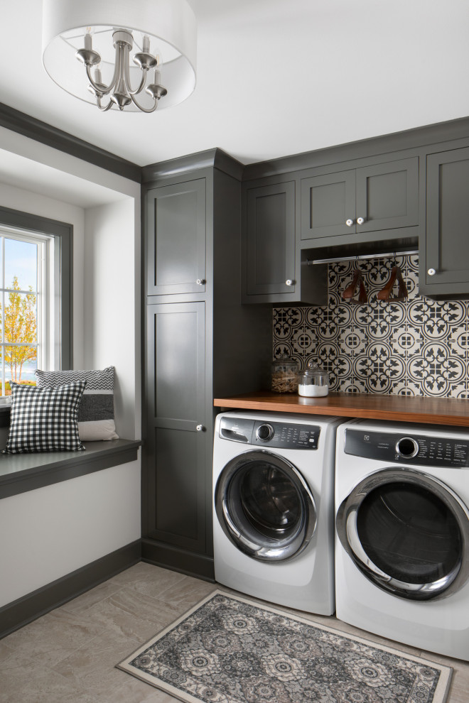 This is an example of a transitional laundry room in Milwaukee with grey cabinets, wood benchtops, multi-coloured splashback, ceramic splashback, grey walls, ceramic floors, a side-by-side washer and dryer, grey floor and brown benchtop.