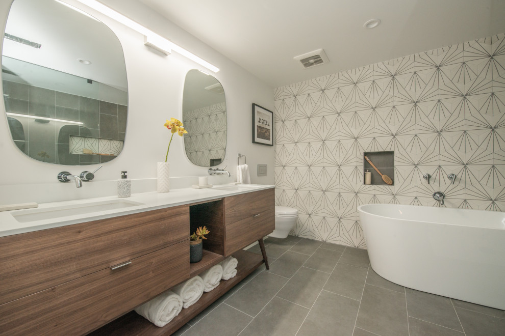 Large retro ensuite bathroom in Los Angeles with freestanding cabinets, medium wood cabinets, a freestanding bath, a built-in shower, a wall mounted toilet, grey tiles, cement tiles, white walls, porcelain flooring, a submerged sink, engineered stone worktops, grey floors, an open shower, white worktops, a wall niche, double sinks and a freestanding vanity unit.