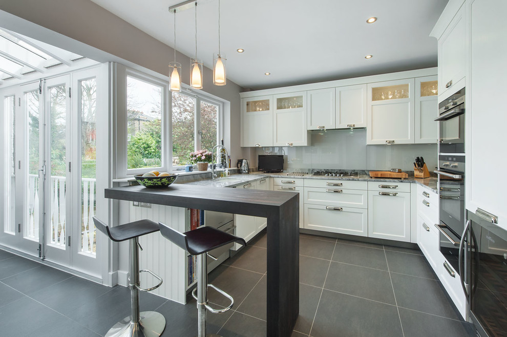 Design ideas for a mid-sized transitional kitchen in London with shaker cabinets, white cabinets, quartzite benchtops, grey splashback, glass sheet splashback, black appliances, porcelain floors, a peninsula and an undermount sink.