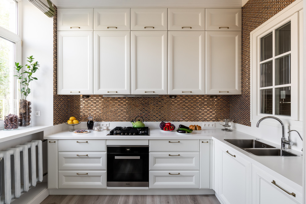 Mid-sized scandinavian l-shaped separate kitchen in Moscow with white cabinets, solid surface benchtops, mosaic tile splashback, stainless steel appliances, laminate floors, white benchtop, a drop-in sink, recessed-panel cabinets, brown splashback and grey floor.