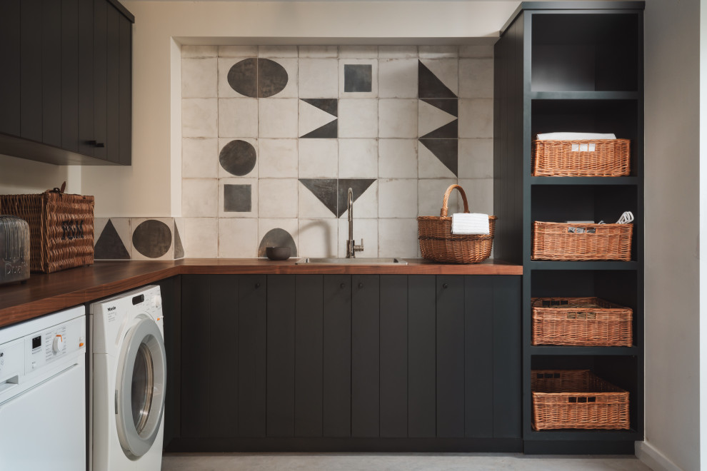 Photo of a country l-shaped dedicated laundry room in Other with a drop-in sink, flat-panel cabinets, black cabinets, wood benchtops, grey walls, a side-by-side washer and dryer, grey floor and brown benchtop.