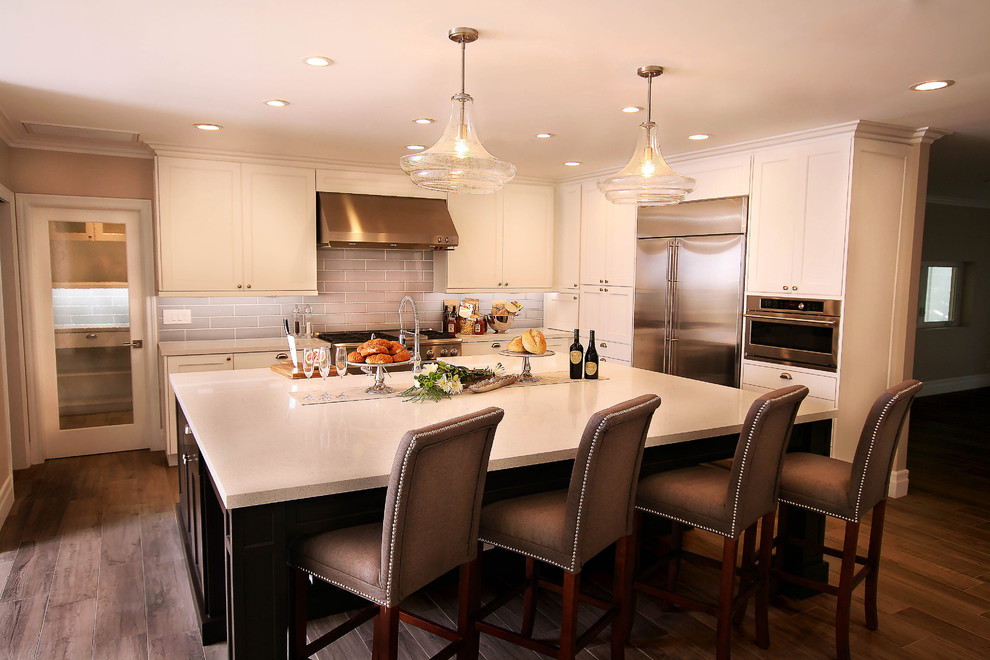 Example of a large transitional u-shaped light wood floor kitchen pantry design with a farmhouse sink, shaker cabinets, white cabinets, quartz countertops, gray backsplash, glass tile backsplash, stainless steel appliances and an island